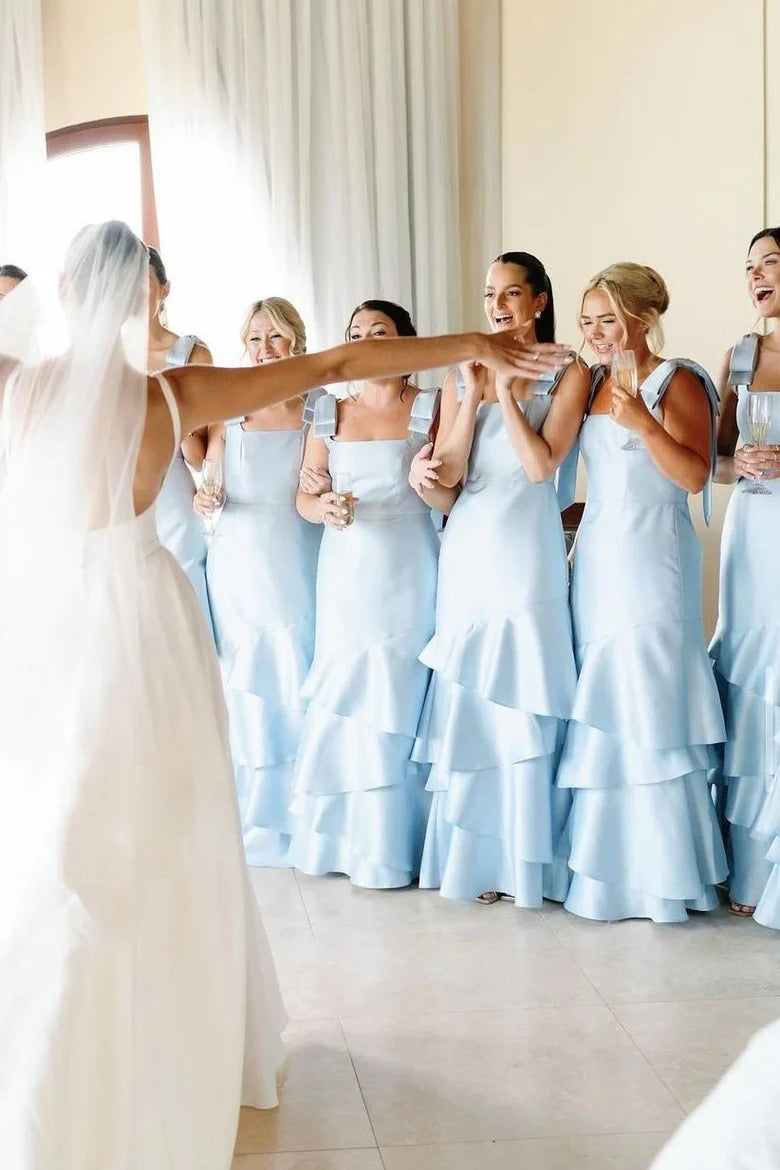 Model wearing a light blue mermaid bridesmaid dress with delicate straps, bows, and a tiered satin skirt.